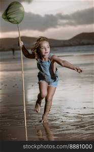 Full body barefoot girl in denim overall shorts with scoop net pointing away and running near sea at sundown on Famara Beach in Lanzarote, Spain. Barefoot girl with scoop net running on beach