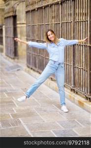 Full body cheerful adult female in casual clothes raising arm and leg and looking at camera with smile, while grasping bar of aged fence on city street. Playful woman grasping old fence
