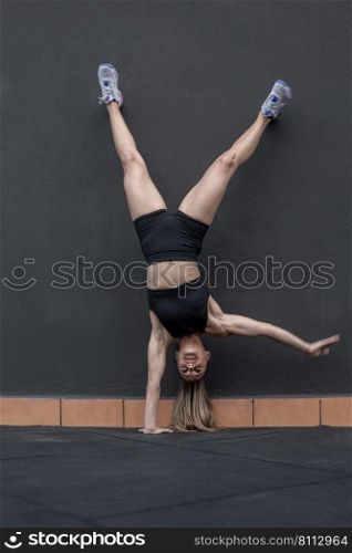 Full body female athlete in sportswear doing one arm handstand near wall during intense fitness training in gym. Sportswoman doing one arm handstand