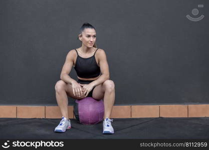 Full body female athlete in sportswear sitting on medicine ball and looking away against black wall during break in fitness training in gym. Sportswoman taking break in workout
