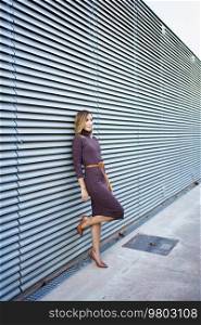 Full body female entrepreneur in stylish dress and high heeled shoes leaning on metal wall of modern building on city street. Businesswoman leaning on wall