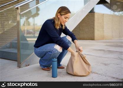 Full body female in stylish clothes sitting on haunches near steps and searching for keys inside bag outside modern apartment building. Woman searching for keys in bag