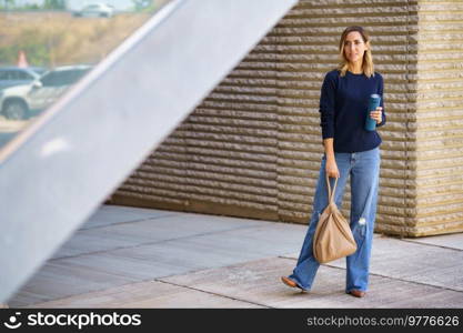 Full body female in stylish jeans and sweater with bag and thermos looking away while standing on pavement outside modern building. Stylish woman standing on street