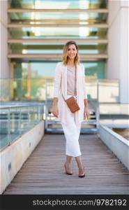 Full body female in stylish outfit with bag and cellphone standing on footbridge outside contemporary building and looking at camera with smile. Stylish woman on footbridge outside modern building