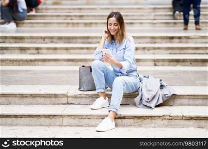 Full body glad adult woman in casual clothes touching hair and watching video on smartphone, while sitting on steps near stylish bag and coat in daytime on city street. Cheerful female watching video on street