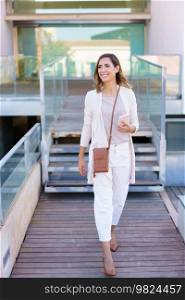 Full body happy adult woman in stylish clothes with smartphone smiling and looking away while walking on path against modern building. Cheerful stylish female walking on boardwalk
