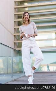 Full body happy female in stylish clothes, opening bottle of water and looking at camera with smile while standing on bridge with glass railing outside contemporary building. Cheerful female with bottle on footbridge