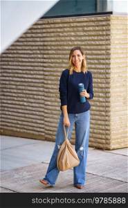Full body happy female in stylish clothes with thermos and bag smiling and looking at camera while standing on city street. Cheerful woman with thermos and bag on street