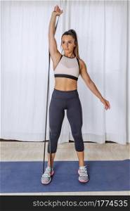 Full body of active fit young female in sportswear doing single arm shoulder press exercise with resistance band during fitness workout at home. Fit woman doing resistance band workout at home