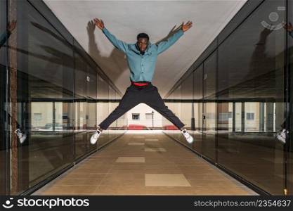 Full body of active young African American guy in trendy outfit and sneakers jumping high with raised arms in passage between glass walls. Trendy serious black man jumping in passage and looking at camera