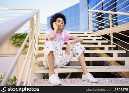 Full body of African American female in casual wear looking away while speaking on cellphone on stairs near building in city. Black woman having phone conversation on staircase