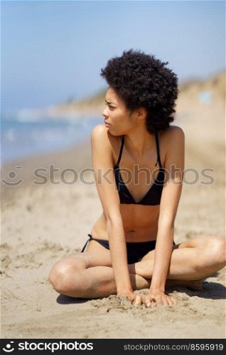 Full body of African American female in swimwear with Afro hair looking into distance while sitting on sandy beach near sea in summer. Black woman in bikini sitting near sea