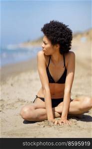 Full body of African American female in swimwear with Afro hair looking into distance while sitting on sandy beach near sea in summer. Black woman in bikini sitting near sea
