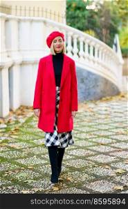 Full body of attractive female in red coat with beret standing on street with closed eyes near white fence in city. Stylish woman in red outfit on street