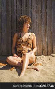 Full body of barefoot female in swimwear and knitted beach dress sitting on sand leaning on wooden fence looking away thoughtfully during sunny day. Sensual woman in crochet beach cover up sitting on sand