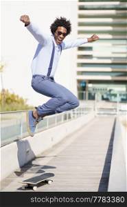 Full body of carefree African American male in smart casual style jumping above skateboard in city and looking at camera. Cheerful black man riding skateboard and jumping