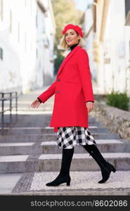 Full body of charming female in red beret looking at camera with smile while strolling on stone stairs in city. Stylish woman in coat walking on steps