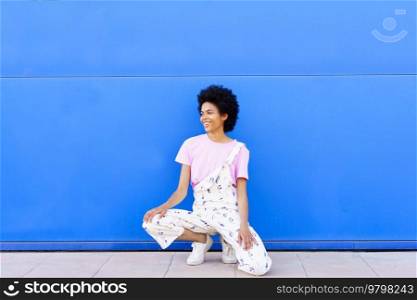 Full body of cheerful African American female in overall looking away with smile while sitting on haunches against blue background. Content black woman hunkering down near wall
