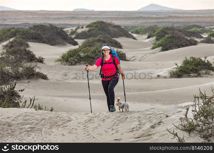 Full body of cheerful female hiker with trekking poles and Yorkshire Terrier dog hiking on Famara beach in summer and looking at camera. Happy traveling woman with dog on sandy terrain