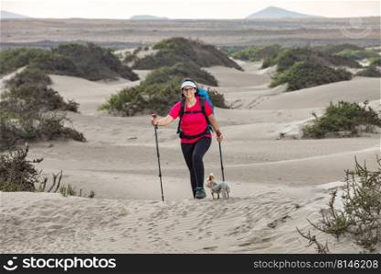 Full body of cheerful female hiker with trekking poles and Yorkshire Terrier dog hiking on Famara beach in summer and looking at camera. Happy traveling woman with dog on sandy terrain