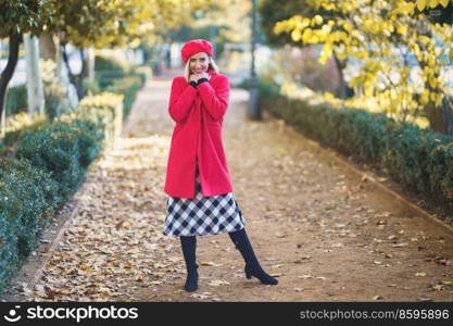 Full body of cheerful female in red coat and beret looking at camera while standing on pathway in park on autumn day. Fashionable woman in autumn park
