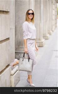 Full body of confident stylish woman gray outfit and sunglasses standing near old building with raised leg and holding handbag at city street. Stylish business woman with handbag standing near building