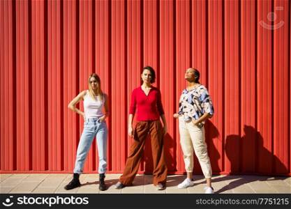 Full body of confident young multiracial female models in casual clothes standing near red metallic wall on sunny day in city. Stylish young diverse ladies standing against red fence on street