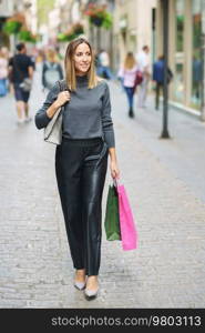 Full body of content adult woman in leather pants and heels carrying colorful paper bags after shopping and strolling on cobblestone town street smiling away. Stylish lady with shopping bags walking on street
