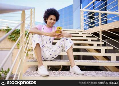 Full body of content African American female in casual wear texting message on cellphone while sitting on stairway in city. Black woman browsing smartphone on staircase