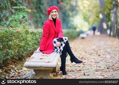 Full body of elegant female in stylish autumn outfit sitting on stone bench in autumn park covered with fade fallen leaves and smiling at camera. Smiling woman sitting on bench in autumn park