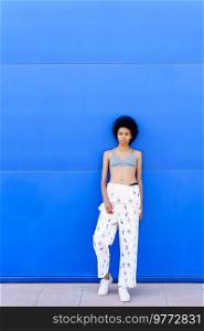 Full body of fit African American female in bra and overall looking away while standing on blue background in city. Black woman in bra near blue wall