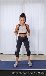 Full body of focused young athletic female in activewear doing exercise with resistance band during intense functional workout at home. Fit woman training muscles with elastic band
