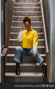 Full body of modern young black female in trendy yellow shirt and sunglasses sitting on staircase and looking at camera. Fashionable ethnic hipster woman in yellow outfit