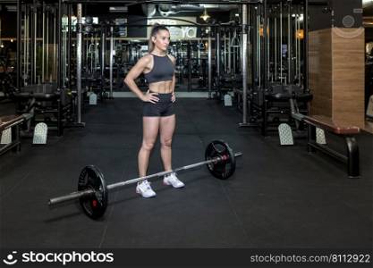 Full body of motivated young muscular female athlete with long ponytail in sportswear standing with hands on waist and looking away before lifting heavy barbell in gym. Fit young sportswoman standing near barbell before weightlifting exercise
