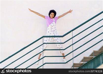 Full body of optimistic African American female in overall looking at camera while star jumping on staircase against white background. Cheerful black woman jumping on stairway