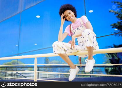 Full body of optimistic African American female looking at camera while sitting on metal fence near glass building in city. Cheerful black woman sitting on railing