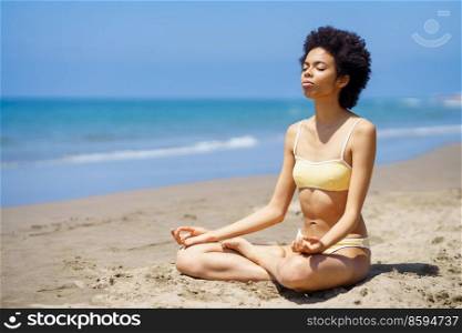 Full body of peaceful slim black female tourist in bikini sitting in Lotus pose on sandy seashore with closed eyes, while meditating during yoga session on sunny summer day. Fit young African American woman in Padmasana meditating on sandy beach