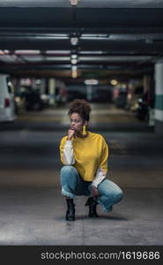 Full body of pensive young African American female in trendy casual outfit and earrings squatting and looking away thoughtfully on underground parking. Stylish hipster black woman on underground parking