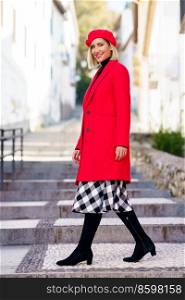 Full body of positive female in red coat and beret looking at camera while walking on stairs near buildings in city. Fashionable woman on steps in city