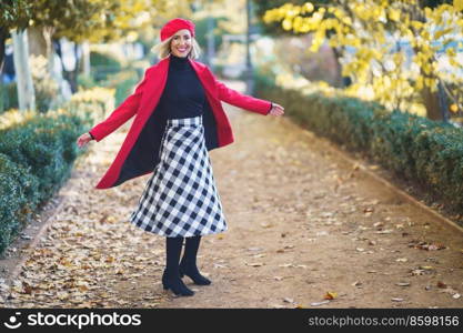 Full body of positive female in red outfit looking at camera while spinning around on pathway in autumn park with trees. Stylish woman spinning around in park