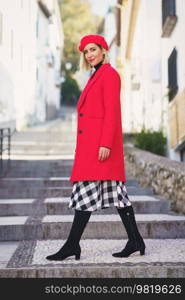 Full body of positive female in stylish red outfit looking away while walking on stone steps on streets with buildings. Stylish woman on stairs in city