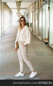 Full body of positive young female in beige blouse and pants with shoulder bag looking at camera smiling. Stylish woman in sunglasses with hand in pocket