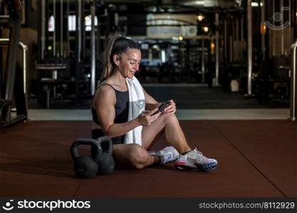 Full body of positive young muscular female athlete with long hair in sportswear and towel on shoulder smiling while messaging on smartphone sitting on floor near kettlebells after workout. Smiling sportswoman using smartphone while resting after training in gym