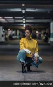 Full body of serious millennial African American female in modern trendy yellow shirt and jeans hunkering down and looking away in underground parking lot. Young black woman in bright trendy outfit squatting on parking lot