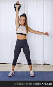 Full body of strong fit young female in sportswear performing strength exercise with kettlebell in raised hand while training alone at home. Determined fit woman training with kettlebell at home