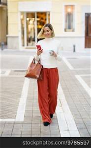 Full body of stylish female carrying handbag and cup of takeaway coffee while walking on street and using cellphone. Smiling woman with takeaway coffee and bag browsing smartphone
