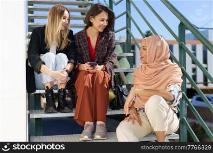 Full body of trendy young multiethnic women friends in stylish outfits sitting on metal stairs on city street and chatting on sunny day. Company of smiling women sitting on stairs and talking
