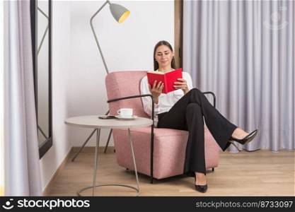 Full body of young elegant female executive manager in classy outfit with long dark hair reading notes while sitting in comfortable armchair near table with coffee cup and smartphone. Confident young classy businesswoman reading notebook sitting in armchair in hotel room