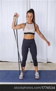Full body of young fit female in sportswear doing one arm bicep curl exercise with resistance rope during fitness workout at home. Active woman doing bicep exercise with resistance band