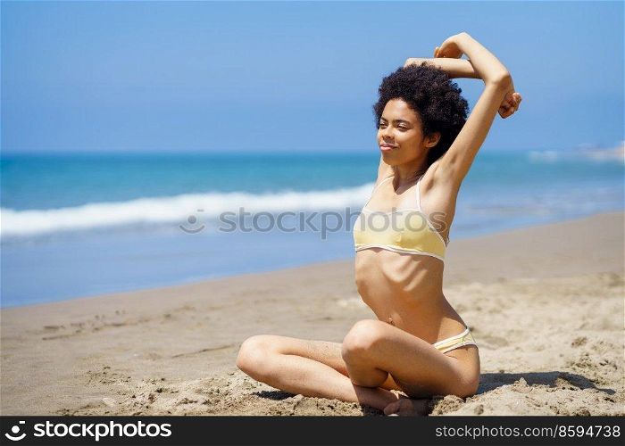 Full body of young positive African American female millennial with curly dark hair in trendy bikini, stretching arms while warming up on sandy coast against cloudless blue sky and sea. Smiling black lady stretching arms on sandy beach during summer vacation